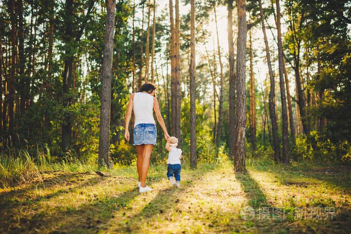 美丽而时髦的母亲在公园里牵着一岁的女孩的手, 在高大的针叶树的背景
