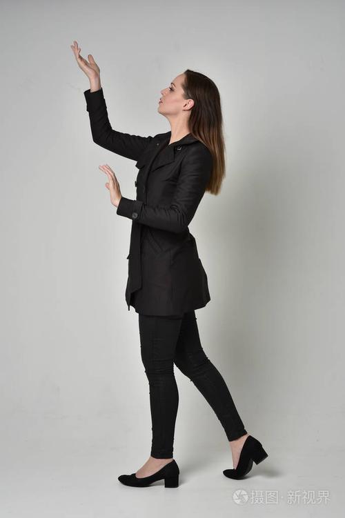 一个黑发女孩穿着长长的黑色外套站在侧面轮廓灰色工作室背景上的完整