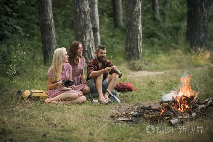 胡子男子和两个姐妹在复古礼服坐在旁边的篝火和烹饪食品户外