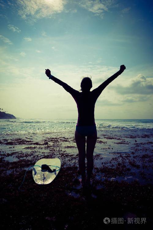 快乐的自由女人冲浪者张开双臂在海边礁的日出时到达天空
