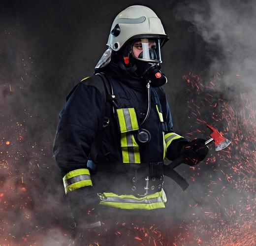 一名身穿制服和氧气面罩的消防员手持一把红色斧头站在火的火花和烟雾