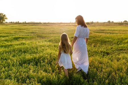 在白色背景下草母亲和女儿牵手走在绿色的草地上, 日落的背景下照片