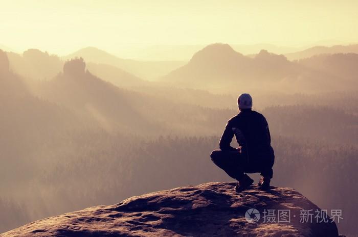 在黑色运动装的年轻男子是坐在悬崖边上,期待迷雾笼罩的山谷贝娄