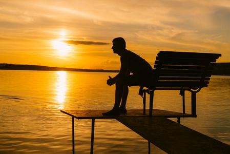 板凳日落成年男子坐在板凳上与折叠的手在河的背景日落照片