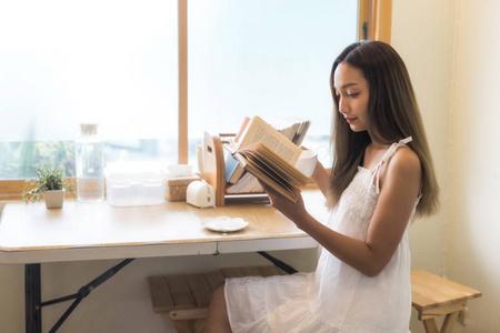 穿着白色连衣裙的亚洲美女坐在窗边看书喝咖啡.