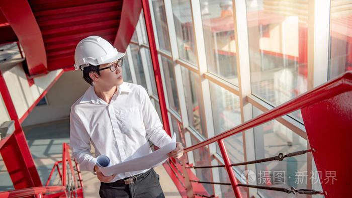 年轻的亚洲男性工程师或建筑师持有蓝图在建筑工地的红色楼梯上佩戴