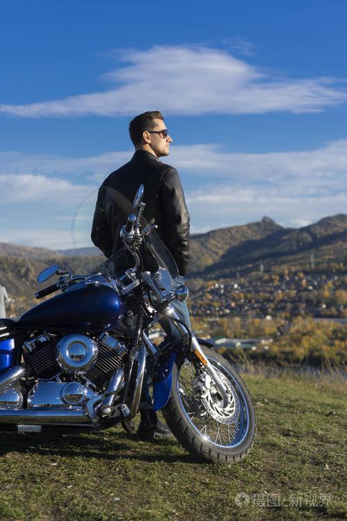 穿着黑色皮夹克太阳镜和牛仔裤的年轻人呆在户外,山上休息的摩托车上.