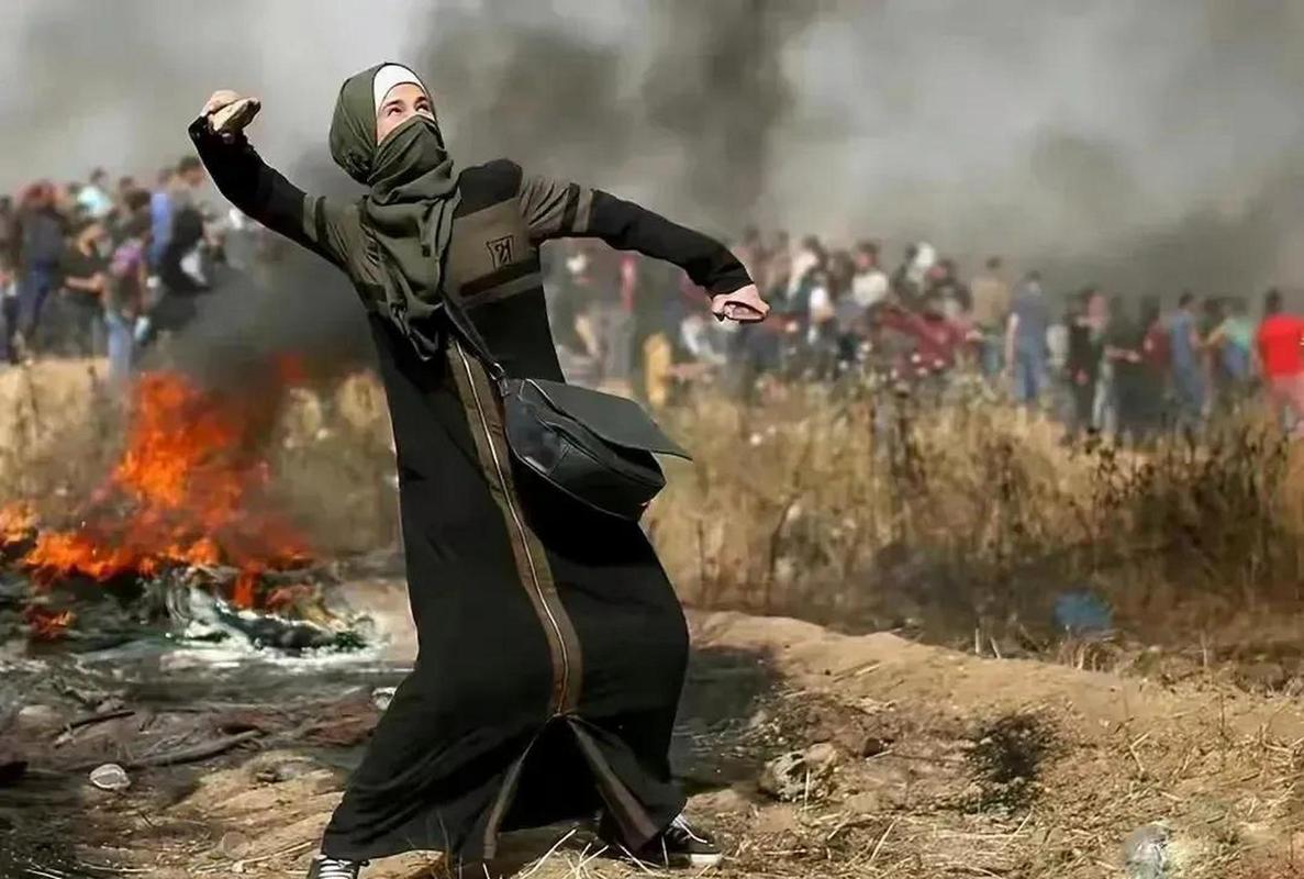 在浓烟滚滚的背景下,一个全身黑袍的巴勒斯坦妇女,站在满目疮痍的土地
