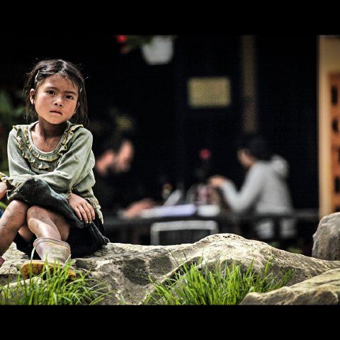 孤独的小女孩,安静地坐立在街道前的一块石头之上,身后的外国人,正在