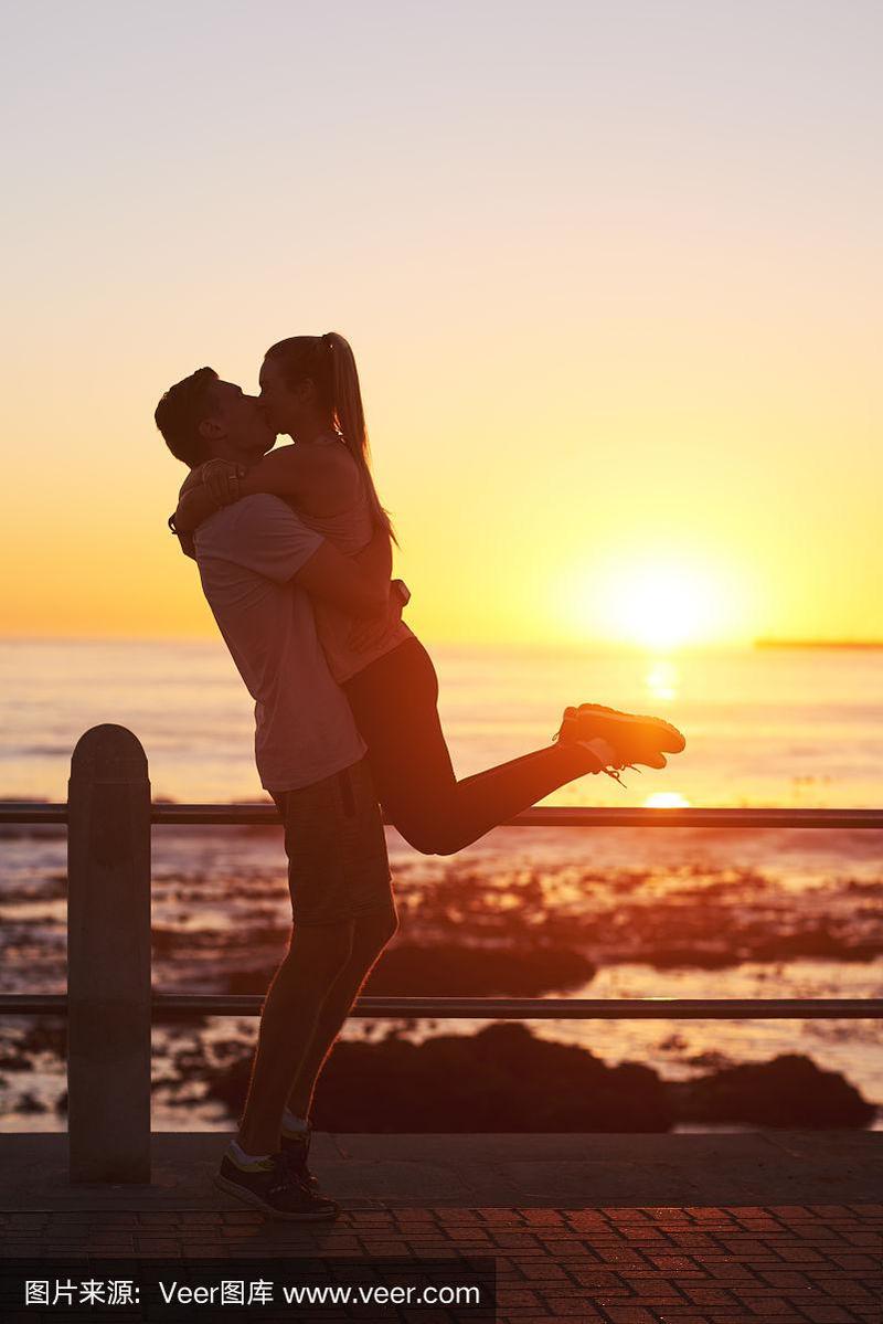 男人的剪影举起女朋友在日落时在海滩上亲吻
