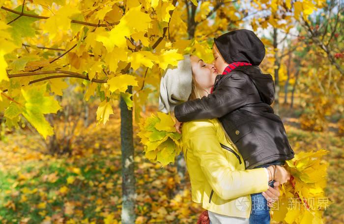 儿子在秋季公园的落叶中亲吻妈妈.水平肖像
