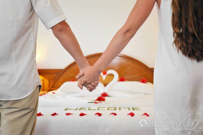 年轻夫妇手拉手在酒店房间与装饰床蜜月理念